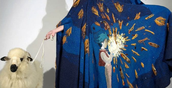 woman poses beside sheep while she wears a dark blue cape and holds up a bag