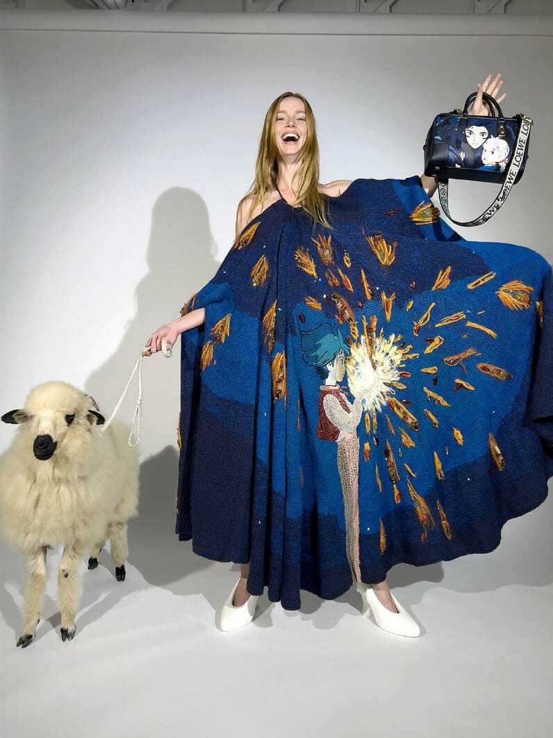 woman poses beside sheep while she wears a dark blue cape and holds up a bag