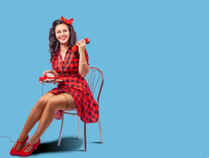 Retro dressed woman models on chair with old style red telephone